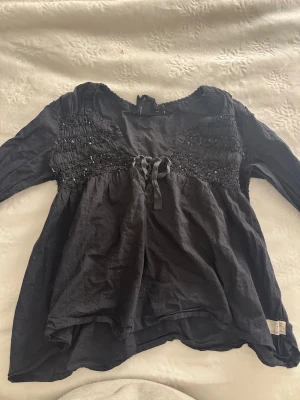 Svart blus med paljettdetaljer - Svart blus med långa ärmar och smockad överdel dekorerad med små svarta paljetter. Blusen har en söt rosett framtill och lätt rynkad nederdel för en avslappnad look. Tillverkad i ett tunt, mjukt tyg som känns skönt mot huden.