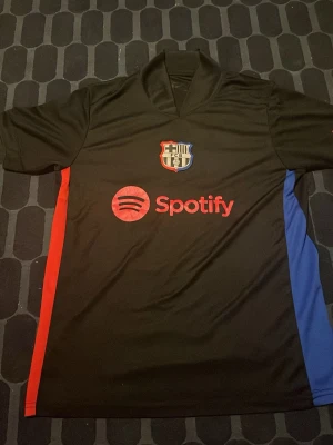 Svart FC Barcelona fotbollströja - Säljer en svart FC Barcelona fotbollströja med klubbmärke på bröstet och röd Spotify-logga framtill. Tröjan har korta ärmar och färgdetaljer i rött och blått på sidorna. Perfekt för dig som älskar fotboll och vill stötta Barca!