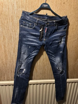 Dsq2 jeans  - Säljer ett par mörkblå jeans med slitningar och patchade detaljer. Jeansen har en unik dragkedjedetalj framtill och klassisk femficksdesign. De är i slim fit-modell och har snygga slitningar på både lår och knän. Perfekta för en trendig streetstil. Midjemått 45cm, längd 105