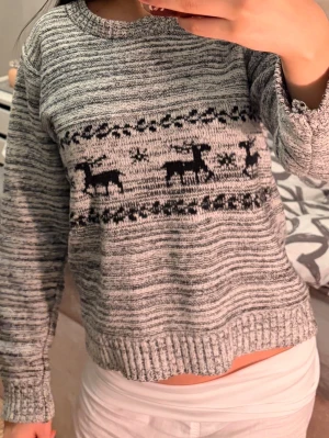 Grå stickad tröja med renmotiv🌸 - Mysig gråmelerad stickad tröja med svart renmotiv och snöflingor över bröstet. Tröjan har rund halsringning, långa ärmar och ribbade muddar vid ärmslut och nederkant. 