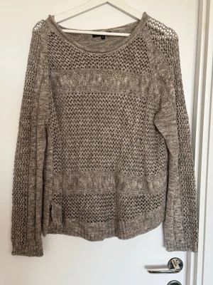Beige/guld stickad tröja med hålmönster - Säljer en beige med gulddetaljer stickad tröja med snyggt hålmönster och rund hals. Tröjan har långa ärmar och ribbade muddar. Perfekt för lager på lager och ger en avslappnad men trendig look.