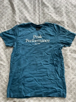 Blå Peak Performance t-shirt - Snygg blå t-shirt från Peak Performance med vit logga och text på bröstet. Klassisk passform med rund hals och korta ärmar. Tillverkad i mjuk bomull som känns skön mot huden. Perfekt för en avslappnad och stilren look.