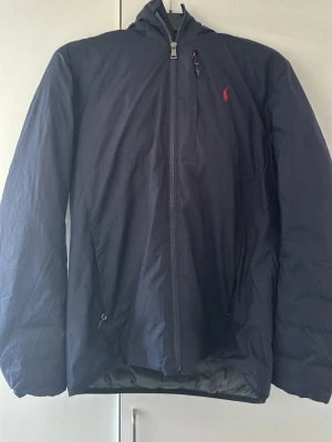 Mörkblå vindjacka Polo Ralph Lauren - Mörkblå jacka från Polo Ralph Lauren med huva och hel dragkedja. Jackan har röd broderad logga på bröstet, två sidofickor och en bröstficka med dragkedja. Tillverkad i lätt och vindtätt material som ger en clean och sportig look. Perfekt för våren och även hösten. Pris kan diskuteras!