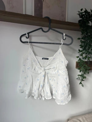 Vit blommig linnetopp GINATRICOT - Söt vit linnetopp från Ginatricot med tunna axelband och små blå blommor. Toppen har en knyt-detalj framtill och är lätt utsvängd nertill för en luftig känsla. Perfekt för varma dagar och enkel att styla med jeans eller kjol.