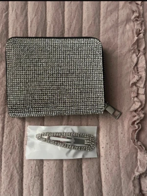 Glittrig silverplånbok med dragkedja - En fyrkantig plånbok täckt med gnistrande silverfärgade strass och dragkedja runtom. Plånboken har flera fack för kort och sedlar. Materialet är syntet med strass på utsidan. Perfekt för dig som gillar att sticka ut och vill ha något extra i väskan.