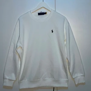 Vit sweatshirt från Polo Ralph Lauren - Klassisk vit sweatshirt från Polo Ralph Lauren med rund halsringning och den ikoniska svarta logobroderingen på bröstet. Tröjan har långa ärmar och ribbade muddar vid ärmslut och nederkant. Perfekt till en clean och stilren look.