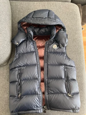 Mörkblå dunväst från Moncler - Snygg mörkblå dunväst från Moncler med avtagbar huva och brunt innerfoder. Västen har två dragkedjefickor framtill och klassisk Moncler–logga på bröstet. Perfekt för lager–på–lager och håller dig varm under kyliga dagar.