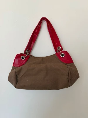 Brun handväska med röda detaljer - Cool brun handväska med glansiga röda handtag och hörn. Väskan har en mjuk form, stora metallöljetter och en dragkedja på baksidan. Perfekt för dig som vill ha en unik accessoar med kontrastfärger och snygga detaljer.