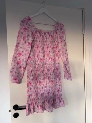 Blommig rosa långärmad klänning - Söt klänning med blommigt mönster i rosa och vitt. Långärmad modell med smockad överdel och volangkant nertill. Klänningen har en rak passform och är tillverkad i ett lätt, mjukt material. Perfekt för vår och sommar.