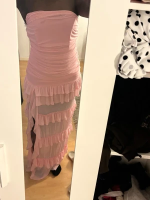 Lång rosa volangklänning omlott - Säljer en lång, axelbandslös rosa klänning med flera lager volanger och transparenta partier. Klänningen har omlottkänsla och är figurnära med en romantisk vibe. 