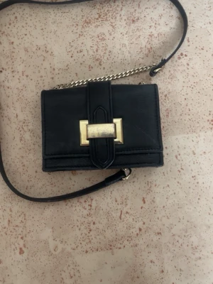 Svart miniväska från DAY ET - Snygg svart miniväska från DAY ET med guldfärgade detaljer och kedja. Väskan har ett stilrent rektangulärt format, dekorativt spänne framtill och justerbar axelrem i både läder och metall. Perfekt för dig som gillar minimalistisk och trendig design.