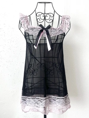 Svart nattlinne med rosett från Music Legs Lingerie - Söt och feminin svart body i transparent mesh med ljuslila spetsdetaljer vid axlar och nederkant. Passar storlek 32-34. Dockans storlek: 34.