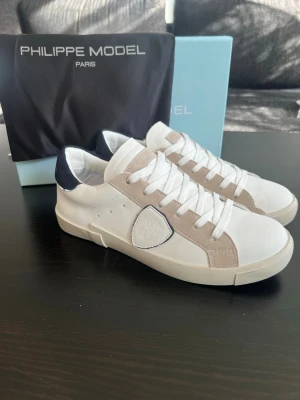 Philippe Model vita sneakers - Snygga vita sneakers från Philippe Model med beige mockadetaljer och mörkblå häl. Klassisk låg modell med vita skosnören och gummisula. Märkets logga på sidan i vitt läder. Perfekta för en clean och stilren look.