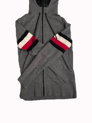 Grå stickad hoodie från Tommy Hilfiger - Säljer en mycket sällsynt grå stickad hoodie med dragkedja och huva i dun material. Ärmarna har breda ränder i svart, vitt och rött som ger en sportig vibe. Tröjan har ribbade muddar och nederkant samt snörning i huvan. Perfekt för dig som gillar en avslappnad men snygg stil. Det står på produkten att storleken är M men den sitter mer som S.
