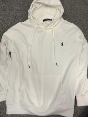 Vit hoodie från Polo Ralph Lauren - Snygg vit hoodie från Polo Ralph Lauren med klassisk huva och dragsnören. Ikoniska loggan broderad på bröstet. Hoodien har en stor magficka och är tillverkad i mjukt material som känns skönt mot huden. Perfekt för en avslappnad och stilren look.