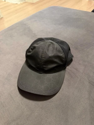 Gucci Keps Nylon - Snygg svart keps från Gucci med diskret GG-mönster över hela framsidan. Kepsen har meshpaneler på sidorna och bak för extra ventilation och en justerbar rem baktill. Perfekt för dig som vill ha en stilren och exklusiv accessoar. Självklart äkta! 