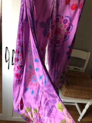 Desigual byxor i lila med mönster stl M - Säljer ett par oanvända långa, lila långbyxor med blommigt mönster från Desigual och smock i midjan. Byxorna är gjorda i tunn bomull så mycket sköna att bära. De har hög slits i yttersidorna. Stl M. Se matchande tunika i annan annons!