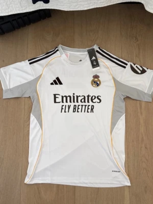 Real Madrid matchtröja Adidas vit - Snygg vit Real Madrid matchtröja från Adidas med svarta detaljer och gula linjer. Tröjan har klubbmärke, Adidas-logga och 'Emirates Fly Better' tryckt på bröstet. Tillverkad i lätt och ventilerande material, perfekt för fotboll eller träning.