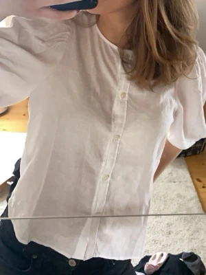 Vit puffärmad linneblus med knappar - Säljer en vit linneblus med puffiga korta ärmar och knäppning framtill. Blusen har en rak passform och är tillverkad i ett tunt, luftigt tyg som känns perfekt för sommaren. Enkel och stilren design som passar till mycket.