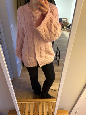 Ljusrosa kabelstickad kofta - Mysig ljusrosa kofta med kabelstickat mönster och stora vita knappar framtill från Gina. Använd fåtal gånger.