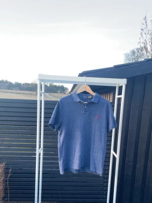 Blå pikétröja från Polo Ralph Lauren - Klassisk blå pikétröja från Polo Ralph Lauren med korta ärmar och krage. Tröjan har knappar vid halsen och den ikoniska röda logotypen broderad på bröstet. Tillverkad i mjuk bomull för en bekväm känsla.