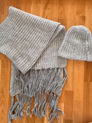 Stickad halsduk med fransar, grå och grön + mössa - paket  - Mysig stickad halsduk i ljusgrått och grön garn med långa fransar i ändarna. Halsduken har en bred, ribbstickad design som ger en chunky och trendig look. Perfekt för att hålla värmen under kalla dagar och passar till de flesta outfits.