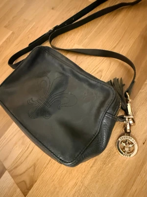 Svart axelväska i äkta läder - Morris Lady - Stilren svart axelväska i slätt äkta läder från Morris Lady med enkel design och justerbar axelrem. Väskan har en klassisk rektangulär form och diskreta metalldetaljer. Perfekt för dig som gillar minimalistisk stil och vill ha plats för det viktigaste.