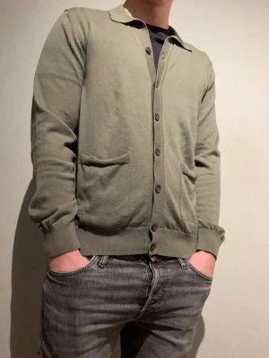 Olivgrön cardigan med knappar - Snygg olivgrön cardigan med knäppning framtill och två fickor. Tröjan har ribbade muddar vid ärmslut och nederkant samt klassisk krage. Perfekt att slänga över en t-shirt för en avslappnad look.