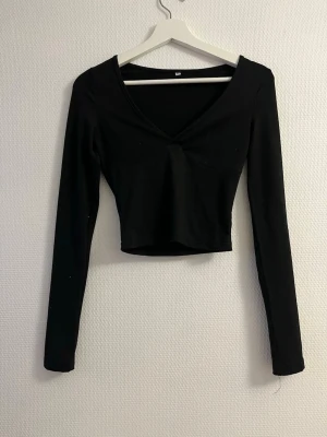 Svart croppad långärmad topp - Snygg svart croppad topp med lång ärm och v-ringad hals. Enkel och stilren design som passar perfekt till jeans eller kjol. Tillverkad i mjukt stretchigt material för en bekväm passform.