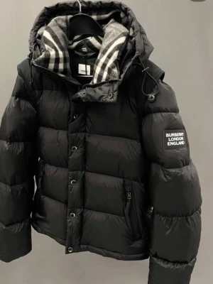 Svart pufferjacka från Burberry - Snygg svart pufferjacka från Burberry med klassiskt rutigt foder i huvan och patch med texten 'Burberry London England' på ärmen. Jackan har avdragbara armar.