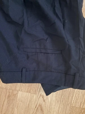 Svarta kostymbyxor med raka ben - Säljer ett par klassiska svarta kostymbyxor med raka ben och hällor för skärp. Byxorna har diskreta fejk bakfickor och är sydda i ett slätt tyg som ger en stilren look. Ganska små i storleken skulle säga att dom är mer storlek 40 än 42.