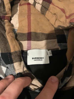 Burberry svart med rutig huva - Säljer en svart skjorta från Burberry med ikonisk beige, svart och röd rutig huva. Skjortan är tillverkad i 100% bomull och har en clean look med soprena linjer och klassiskt rutmönster på huvan. Perfekt för dig som vill ha något stilrent men ändå sticker ut.