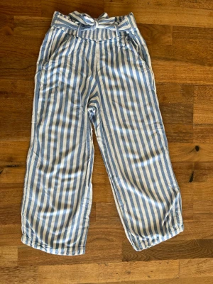 Randiga blåvita linnebyxor med knytband - Säljer ett par luftiga linnebyxor med blå och vita vertikala ränder och ett snyggt knytband i midjan. Byxorna har raka ben och resår baktill för extra komfort. Perfekta för en chill och avslappnad stil.