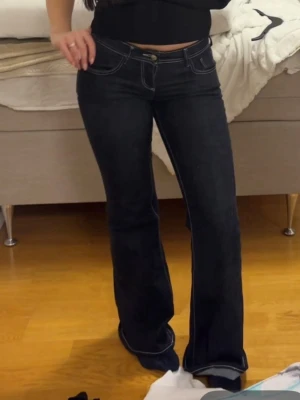 Mörkblå bootcut jeans med kontrastsömmar - Säljer ett par mörkblå bootcut jeans med vita kontrastsömmar och låg midja. Jeansen har klassisk femficksdesign och stängs med knapp och dragkedja. Perfekta för en trendig och avslappnad look.