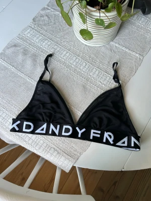 Svart bralette från Frank Dandy - Snygg svart bralette från Frank Dandy med bred resår och vit logga runt om. Triangelformade kupor utan bygel och justerbara axelband. Tillverkad i mjukt och stretchigt mesh. 
