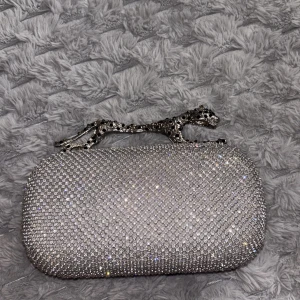 Silver kuvertväska med strass och panter - Säljer en glittrig silverfärgad kuvertväska täckt av strass med ett unikt handtag i form av en panter. Väskan har en oval form och ger riktigt lyxiga vibes. Perfekt accessoar för att sticka ut och addera extra bling till din outfit.