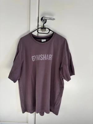 Lila oversized t-shirt från Gymshark - Säljer en lila oversized t-shirt från Gymshark med tryckt logga på bröstet. T-shirten har rund halsringning och korta ärmar. Perfekt för gymmet eller avslappnade dagar. Tillverkad i mjukt bomullsmaterial.