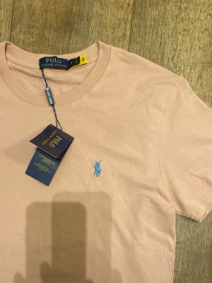 Rosa t-shirt från Polo Ralph Lauren (Helt ny-Oanvänd) - Snygg rosa t-shirt från Ralph Lauren med klassisk rund hals och korta ärmar. T-shirten har den ikoniska blå logotypen broderad på bröstet och är tillverkad i mjuk bomull för en skön känsla. Perfekt till jeans eller shorts. Storlek Medium men passar mer som small. Snabb Leverans!