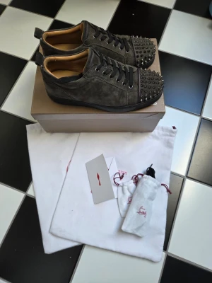 Christian Louboutin gråa sneakers - Gråa sneakers i mocka från Christian Louboutin med svarta skosnören och ikoniska nitar på tån. Låg modell med rundad tå och detaljer i metall. Kommer med originalkartong och dustbags.