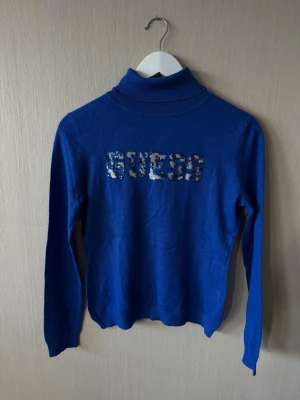 Blå polotopp från Guess med paljetter, S - Blå polotopp från Guess med lång ärm och paljettdetaljer på bröstet som stavar ut loggan. Toppen har ribbad polokrage och är gjord i mjukt material som sitter skönt på kroppen.