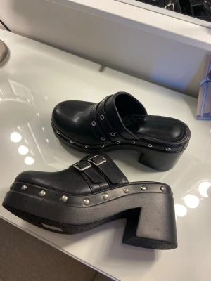 Svarta platåtofflor med nitar och spännen - Säljer ett par svarta platåtofflor med chunky klack, dekorerade med silvriga nitar runt sulan och dubbla spännen över foten. Tofflorna har en cool retrovibe och är tillverkade i konstläder med robust sula. Perfekta för dig som vill sticka ut!