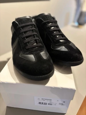 Svarta sneakers från Maison Margiela - Stilrena svarta sneakers från Maison Margiela med ovandel i mocka och läderdetaljer. Klassisk låg modell med svarta skosnören och gummisula med unikt mönster undertill. Perfekta för dig som gillar minimalistisk och clean stil.Skorna är använda 3 gånger och endast lite creasade men det är tyvärr inget man kan råför.priset är inte fast och kan ändras vid snabb affär | För fler frågor och funderingar hör av er.✨