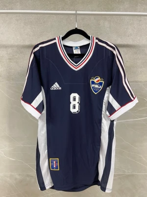 Blå Adidas fotbollströja Mijatovic - Mörkblå fotbollströja från Adidas med vita och röda detaljer, nummer 8 och namnet Mijatovic på ryggen. Tröjan har klubbmärke på bröstet, V-ringad krage och vita sidopaneler. Perfekt för dig som gillar retro fotbollströjor.