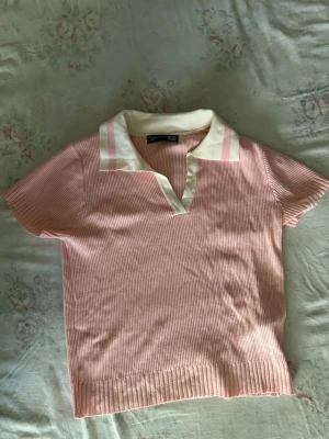 Rosa ribbad pikétröja - Kortärmad rosa ribbad pikétröja  med vit krage och rosa ränder på kragen. Tröjan har v-ringning och är gjord i ett mjukt, stretchigt material som känns bekvämt mot huden. Perfekt för en avslappnad och trendig look.