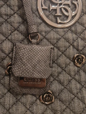 Grå  Guess handväska  - Stilren grå handväska från Guess med quiltat tyg, silverfärgade metallrosor och stort logotyp-emblem. Väskan har en liten accessoar i ormmönstrat konstläder och detaljer i metall. Perfekt för dig som gillar trendiga och unika detaljer.