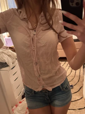 Ljusrosa kortärmad skjorta med knappar - Säljer en ljusrosa skjorta med korta ärmar och knappar framtill. Skjortan har en feminin passform och detaljerade knappar, samt en liten dekorationsdetalj på ärmen med randigt band och knapp. Perfekt till sommaren och enkel att styla till jeans eller kjol.