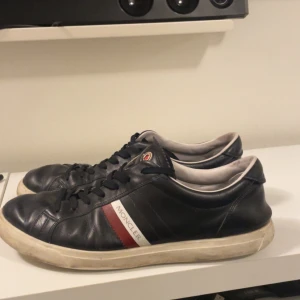 Moncler svarta sneakers i skinn - Svarta sneakers från Moncler med snygga röda och vita detaljer och perfekt för dig som vill ha en bra stil och bra och snygg skor för en billig peng. Säljer skorna så billigt för att det är väl använda och har haft det länge. Det är fortfarande väldigt snygga och värt priset.
