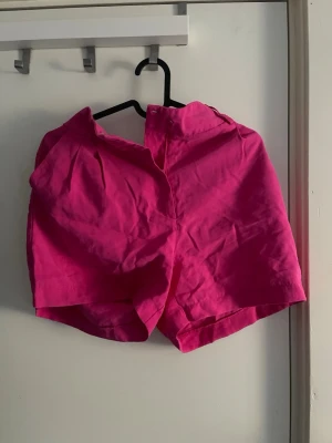 Knallrosa shorts med hög midja - Säljer ett par knallrosa shorts med hög midja och klassisk passform. Shortsen har en enkel design utan synliga detaljer och är tillverkade i ett mjukt bomullsliknande material. Perfekta för varma dagar och ger en färgklick till din outfit.