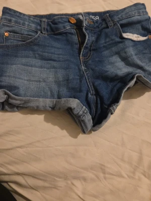 Blå jeansshorts med uppvik - Snygga blå jeansshorts med klassisk femficksdesign och uppvikta benslut. Shortsen har gylf med knapp och dragkedja samt detaljer i kopparfärg. Perfekta för varma dagar och enkel att matcha med olika toppar.