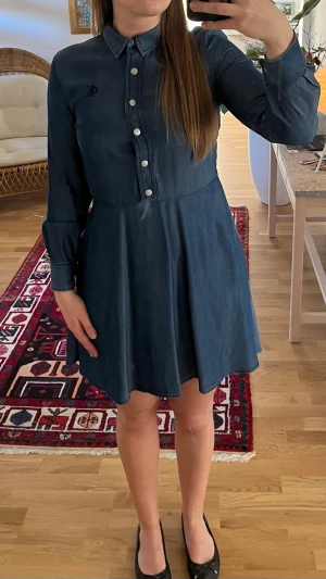 Mörkblå jeansklänning - Snygg mörkblå jeansklänning med klassisk skjortkrage och långa ärmar. Knappslå framtill med vita knappar och lätt utställd kjol. Perfekt för dig som gillar en avslappnad men ändå stilsäker look.